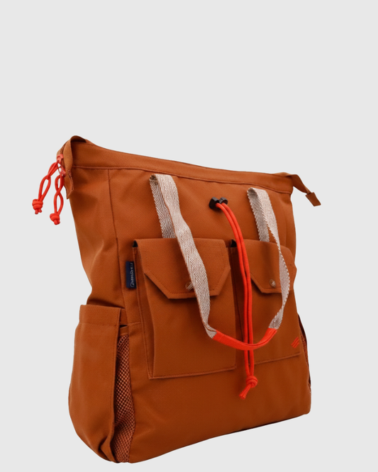 Rustic Caramel (R)evolution™ 27L Utility Convertible Tote (Angle): Burnt orange tote with herringbone handles, red rope pulls, and front flap pockets