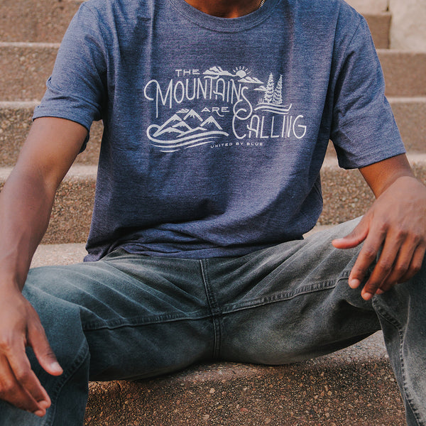 Heathered Navy Mountains Are Calling - Biodegradable Graphic Tee - (All-gender) (In-use): Man sitting on stone steps wearing heathered navy graphic tee outdoors