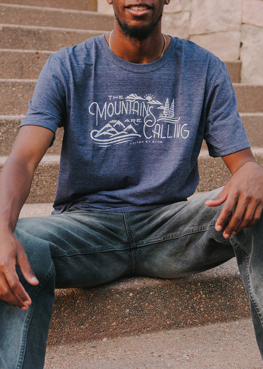 Heathered Navy Mountains Are Calling - Biodegradable Graphic Tee - (All-gender) (In-use): Man sitting on stone steps wearing heathered navy graphic tee outdoors