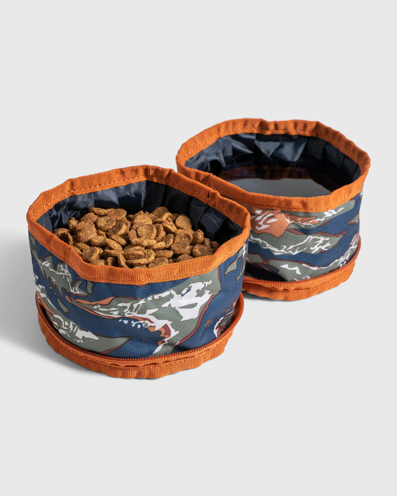 (R)evolution™ Collapsible Double Dog Bowl (Angle): Two collapsible fabric bowls with blue camo print and orange trim, one filled with kibble