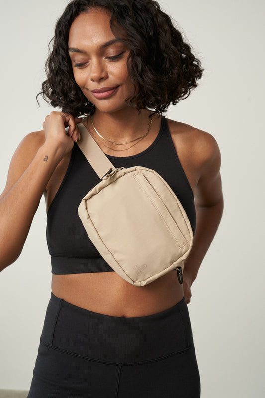 Sand (Re)active™ 2L Convertible Crossbody (In-use): Woman wearing beige crossbody sling bag diagonally over black athletic top