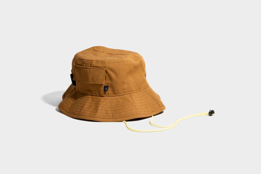 HONEY UTILITY BUCKET HAT (Angle): Tan canvas bucket hat with side pocket, chin cord, and black toggle