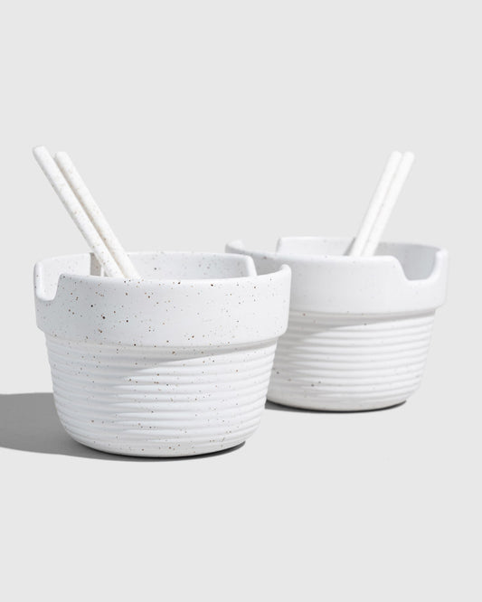 Eggshell Stoneware Stackable Noodle Bowl Set (Angle): Two speckled white ribbed bowls with matching chopsticks resting inside notched rims