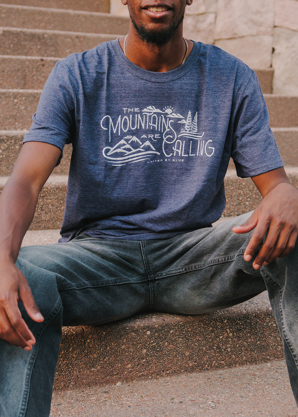 Heathered Navy Mountains Are Calling - Biodegradable Graphic Tee - (All-gender) (In-use): Man sitting on stone steps wearing navy graphic tee with mountain print