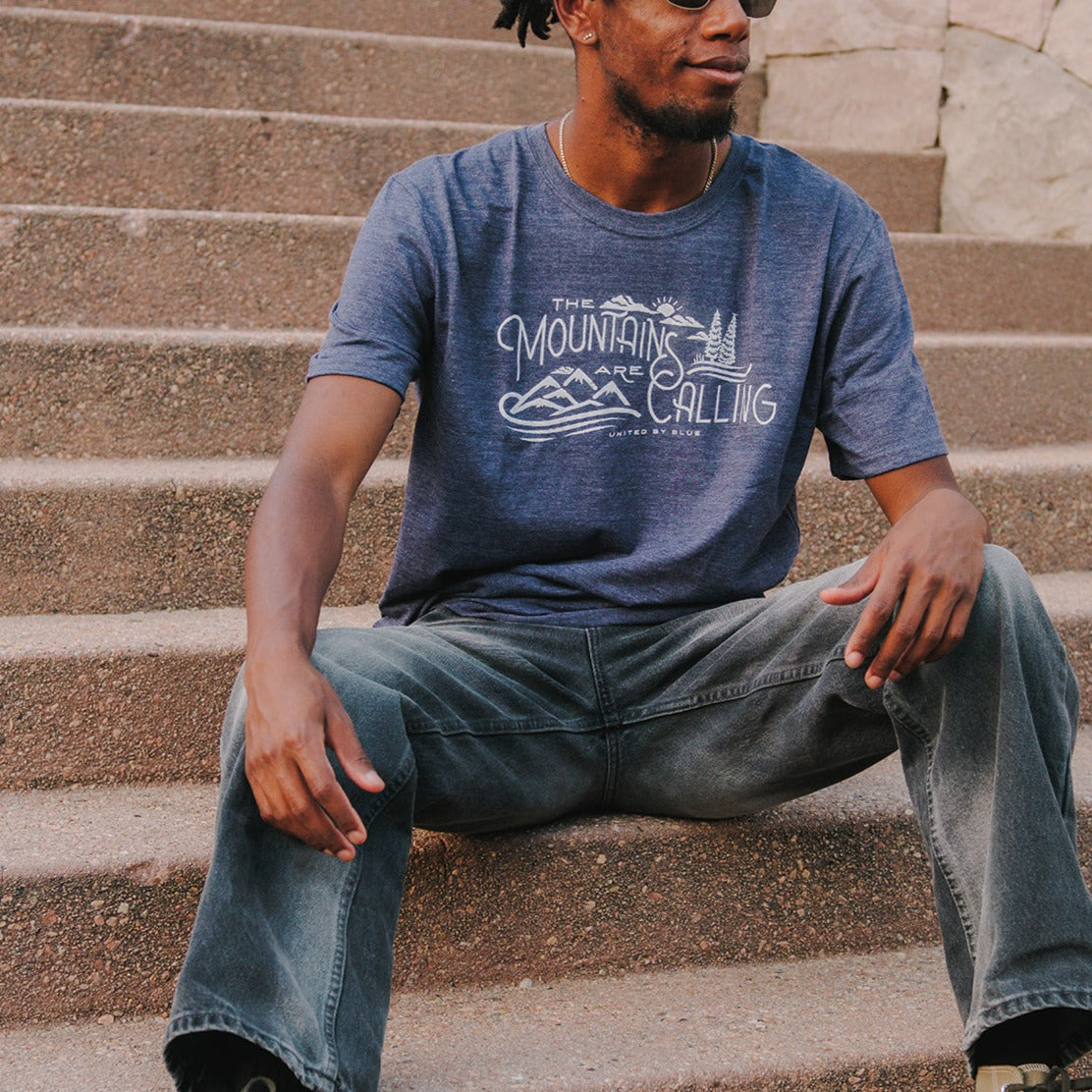 Heathered Navy Mountains Are Calling - Biodegradable Graphic Tee - (All-gender) (In-use): Man seated on stone steps wearing heathered navy tee with mountain graphic