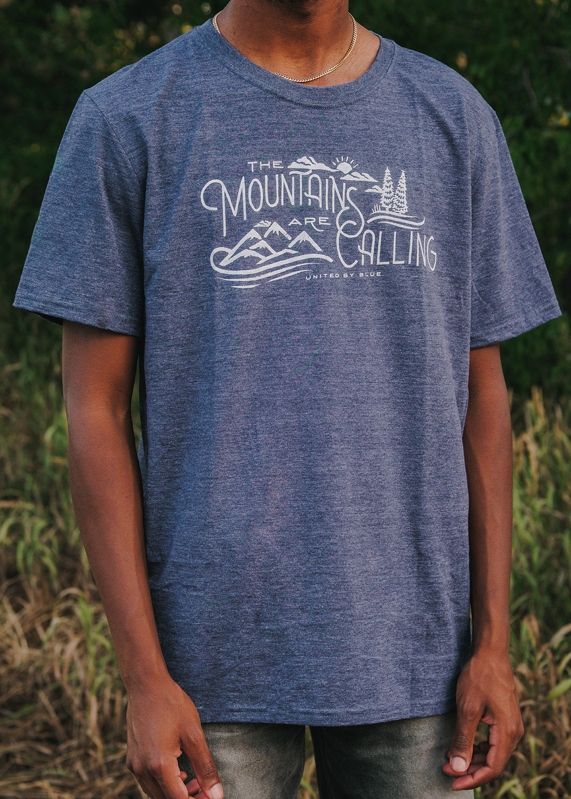 Heathered Navy Mountains Are Calling - Biodegradable Graphic Tee - (All-gender) (In-use): Dark blue heathered tee with white mountain scene graphic, worn outdoors