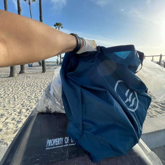 Navy Reusable DIY Cleanup Kit (In-use): Hand holds navy blue bag on beach boardside, palm trees visible