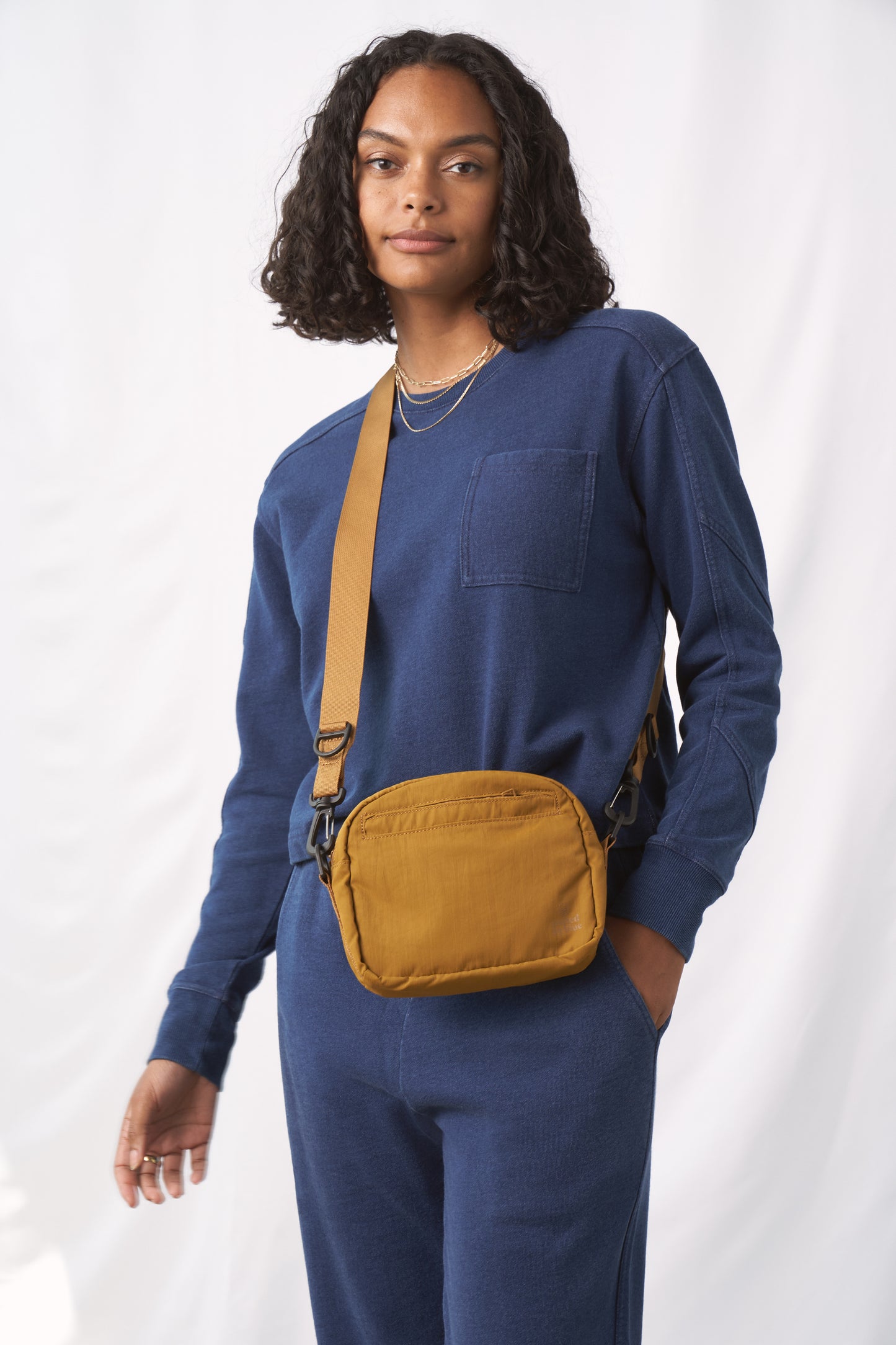 Smoky Ochre (Re)active™ 2L Convertible Crossbody (In-use): Person wearing mustard yellow crossbody bag over blue sweatsuit, crossbody strap