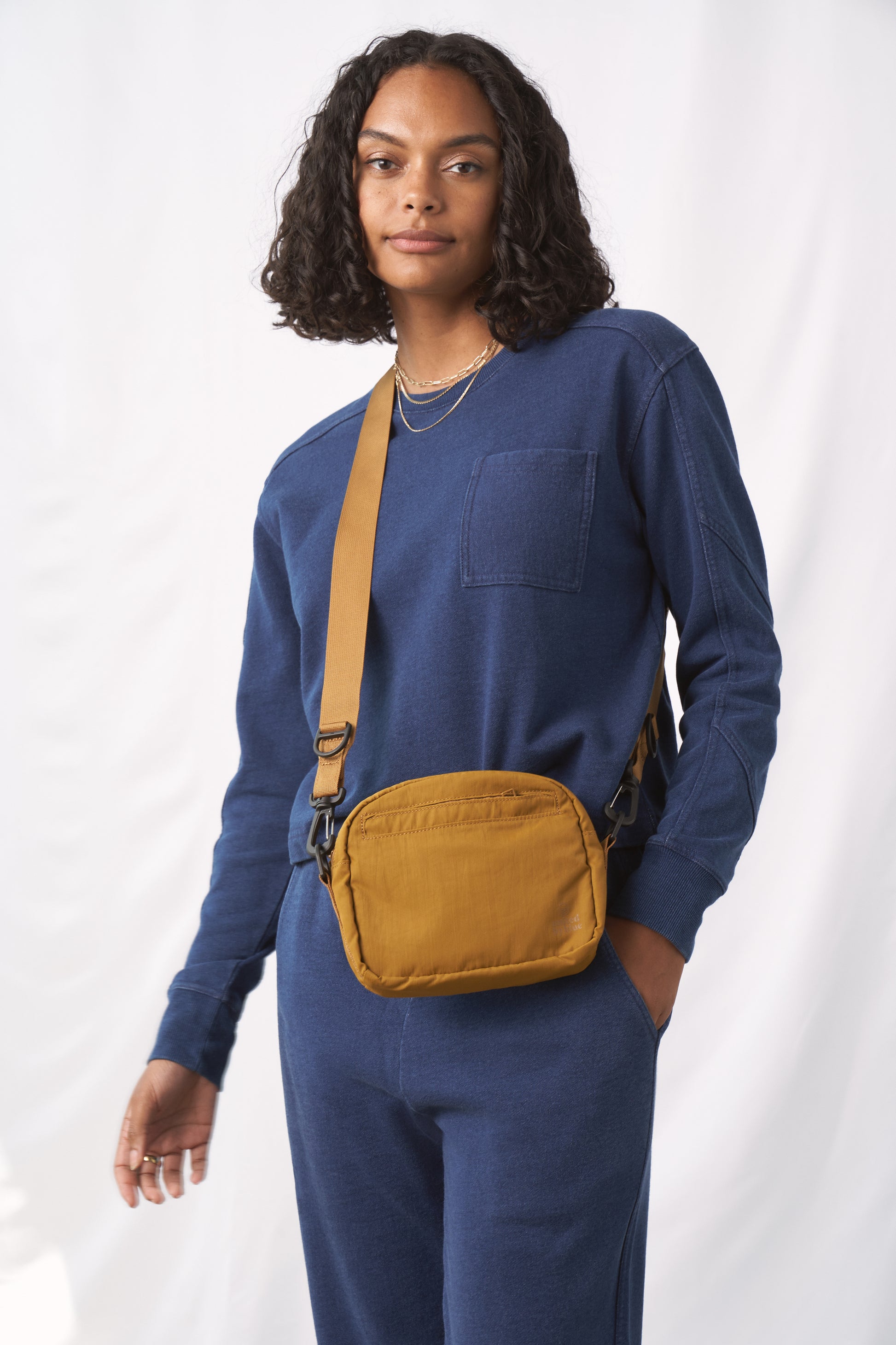 Smoky Ochre (Re)active™ 2L Convertible Crossbody (In-use): Person wearing mustard yellow crossbody bag over blue sweatsuit, crossbody strap