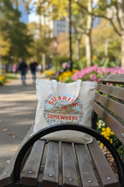It's Always Sunrise Somewhere My Favorite Daily Tote (Lifestyle): Cream canvas tote with mountain sunrise print rests on park bench