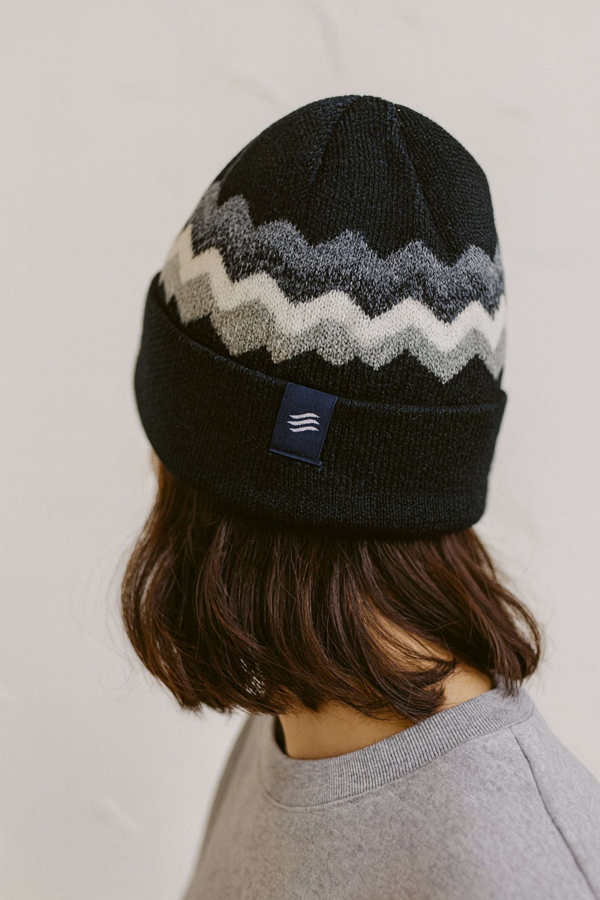 Black Merino Wool Mountain Scape Knit Beanie (In-use): Back view, black beanie with grey white zigzag pattern, small logo patch