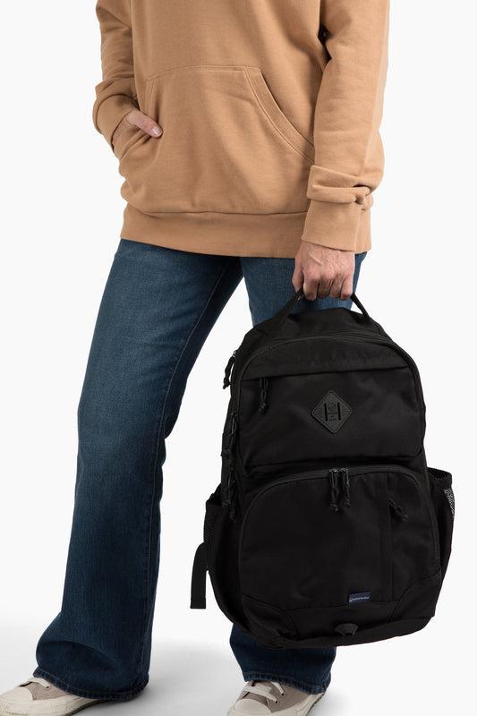 Black (R)evolution™ 25L Transit Pack (In-use): Person in tan hoodie and jeans carrying black backpack at side