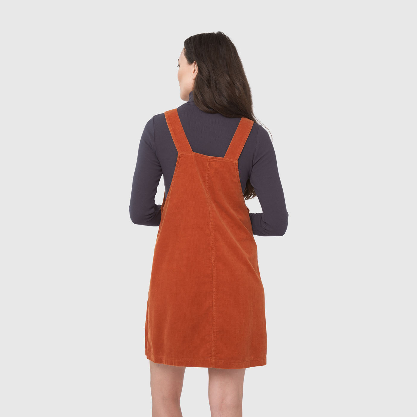 Ginger Corduroy Overall Dress (Back): Rust-orange corduroy pinafore dress with wide straps, worn over navy turtleneck