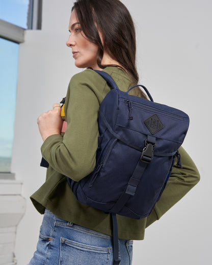 Blue W/ Green (R)evolution™ 9L Sidekick (In-use): Woman wearing navy backpack with flap closure, buckle, and logo patch