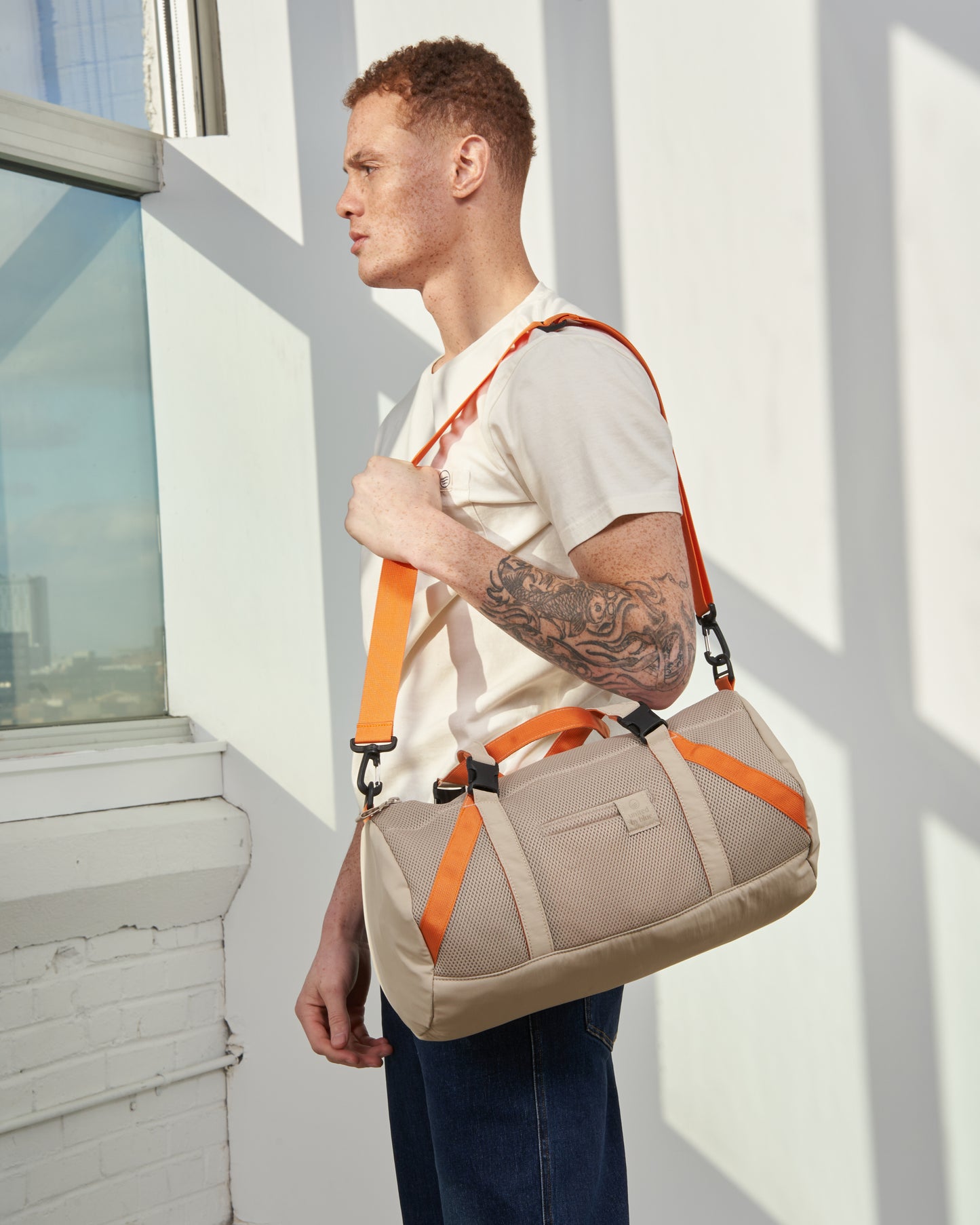 Sand (Re)active™ 16L Mini Duffle (In-use): Man with arm tattoo carries tan duffle with orange straps crossbody