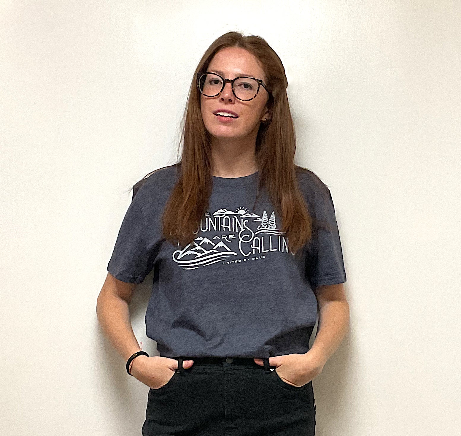 Heathered Navy Mountains Are Calling - Biodegradable Graphic Tee - (All-gender) (In-use): Dark blue tee with white mountain graphic worn by young woman with glasses
