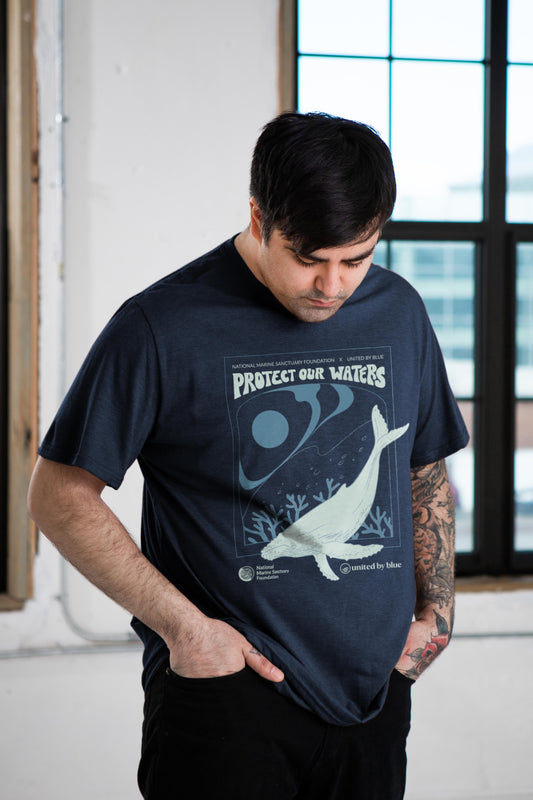 S NMSF x UBB Protect Our Waters - Biodegradable Graphic Tee - (All-gender) (In-use): Man with tattoos wearing navy tee, whale and ocean conservation graphic