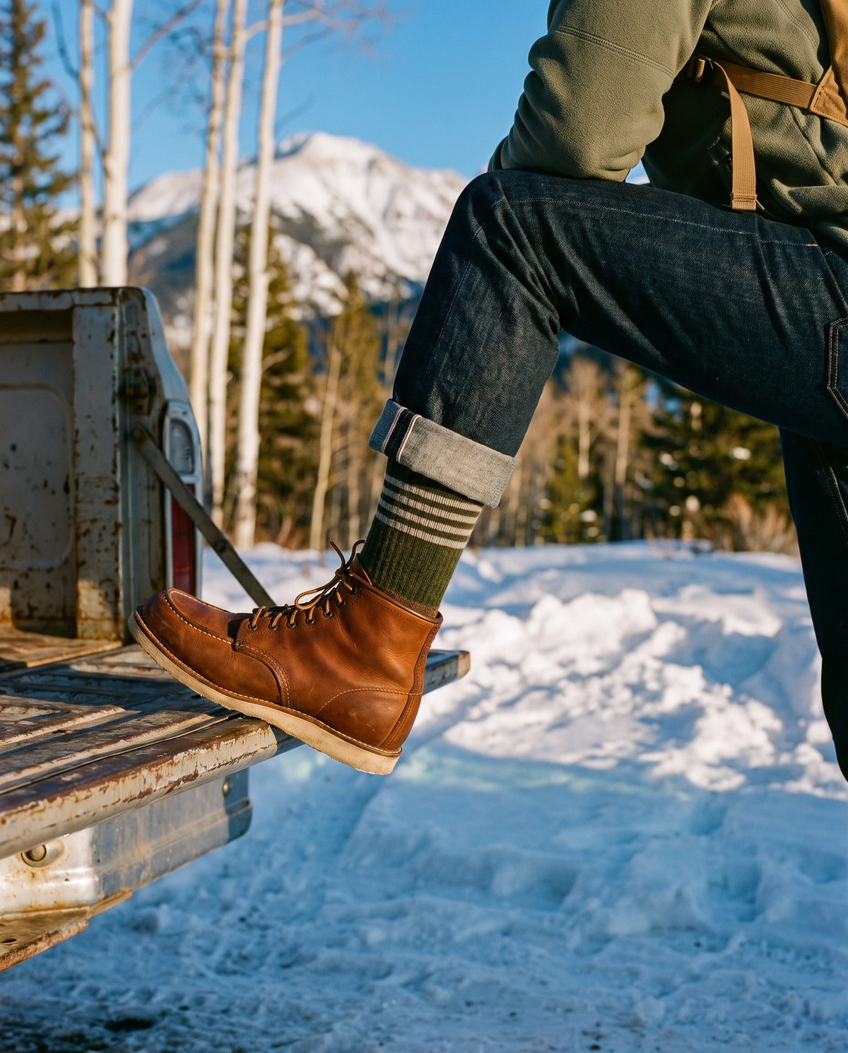 Man in cuffed jeans resting a leather boot on a truck tailgate in Colorado snow, wearing the olive striped Yak Wool Everyday Crew Sock