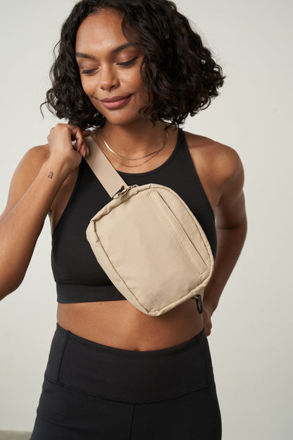 Sand (Re)active™ 2L Convertible Crossbody (In-use): Woman wearing beige crossbody sling bag diagonally over black athletic top