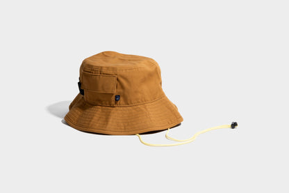 HONEY UTILITY BUCKET HAT (Angle): Tan canvas bucket hat with side pocket, chin cord, and logo patch