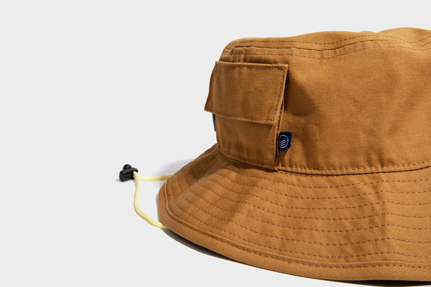 HONEY UTILITY BUCKET HAT (Detail): Close-up of tan bucket hat showing side pocket, chin cord, and logo patch