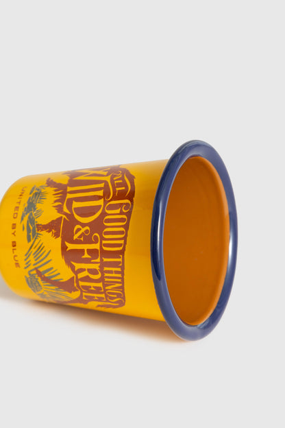 WILD AND FREE WILD AND FREE – 12OZ. ENAMEL TUMBLER (Angle): Yellow enamel tumbler lying on side, blue rim, brown graphic print