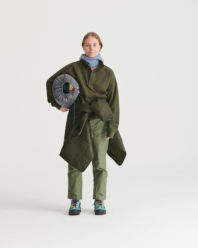 Forest Floor Bison Ultralight Liner Jacket (In-use): Woman wearing olive quilted liner jacket, holding packed sleeping bag, outdoors-ready outfit