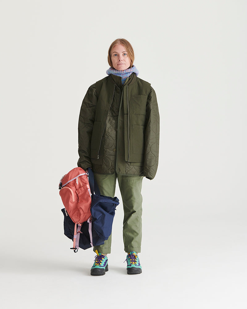 Forest Floor Bison Ultralight Liner Jacket (In-use): Woman standing holding backpacks, wearing olive quilted jacket, matching trousers, colorful boots