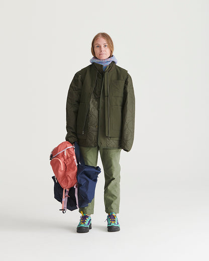 Forest Floor Bison Ultralight Liner Jacket (In-use): Woman standing holding backpacks, wearing olive quilted jacket, matching trousers, colorful boots