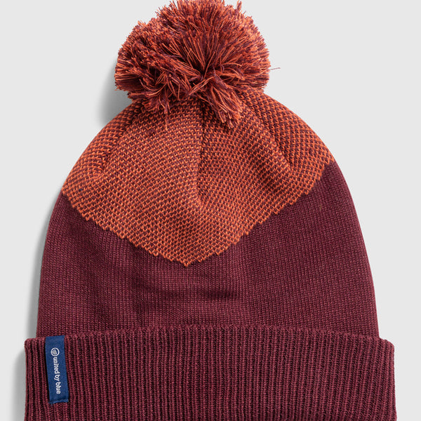 EGGPLANT Recycled Chevron Pom Beanie (Flat-lay): Deep burgundy knit beanie with orange chevron pattern and pom-pom top