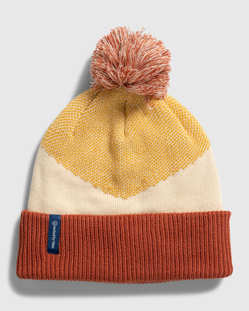 DANDELION Recycled Chevron Pom Beanie (Flat-lay): Rust cuff beanie with cream body, gold chevron pattern, and rust pom-pom