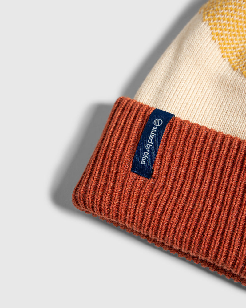 DANDELION Recycled Chevron Pom Beanie (Detail): Close-up of rust cuffed beanie with cream and yellow pattern, blue logo tag
