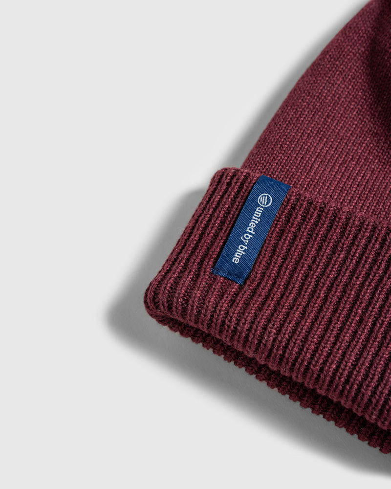 EGGPLANT Recycled Chevron Pom Beanie (Detail): Close-up of dark burgundy ribbed knit cuff with blue brand label