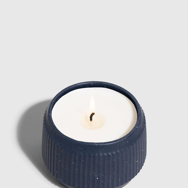 INDIGO 14 oz. Ceramic Keepsake Candle - Surf (Angle): Navy blue ribbed ceramic vessel with white wax candle and lit wick