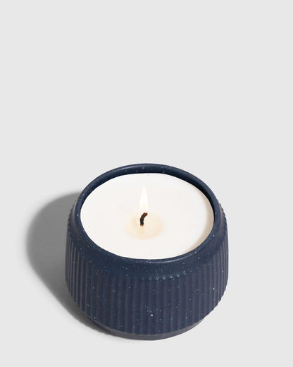 INDIGO 14 oz. Ceramic Keepsake Candle - Surf (Angle): Navy blue ribbed ceramic vessel with white wax candle and lit wick