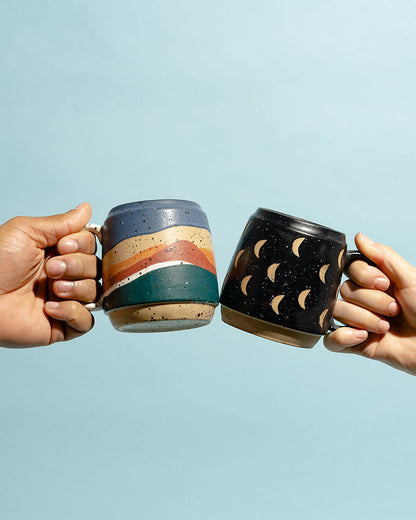 16 oz. Stoneware Mug (In-use): Two hands holding speckled stoneware mugs with rainbow stripes and crescent moon patterns