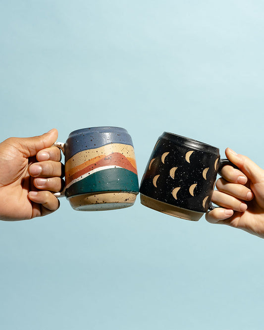 16 oz. Stoneware Mug (In-use): Two hands clinking speckled stoneware mugs, rainbow stripes and crescent moon patterns