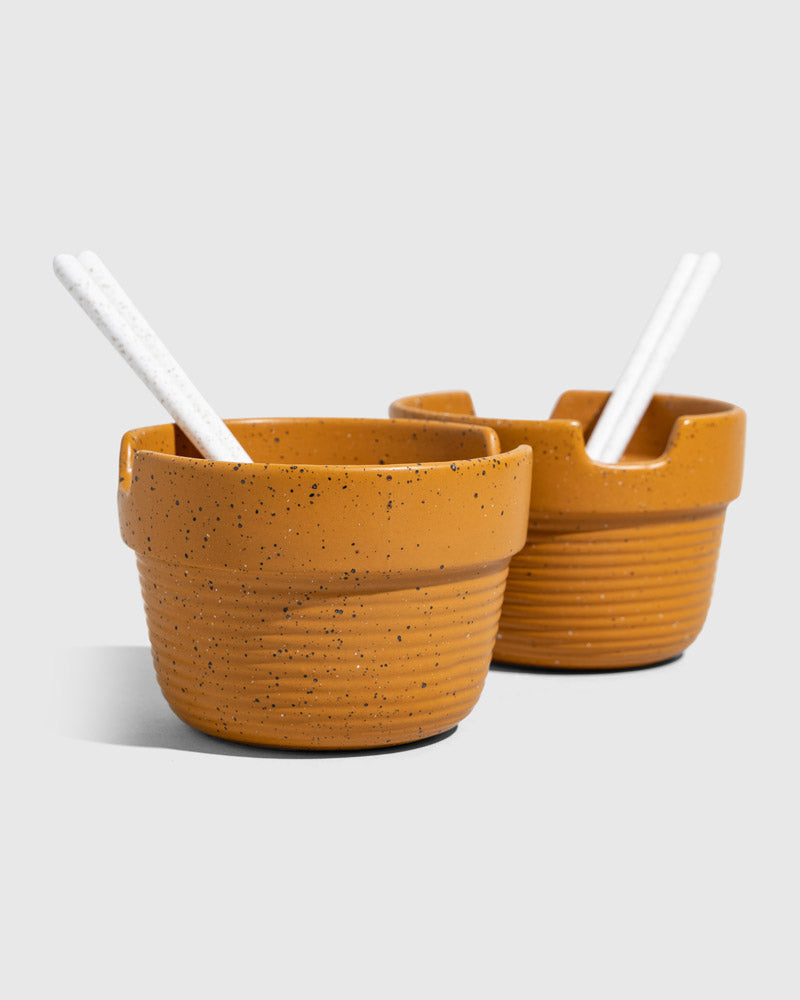 Caramel Stoneware Stackable Noodle Bowl Set (Angle): Two speckled amber ribbed bowls with notched rims and white spoons