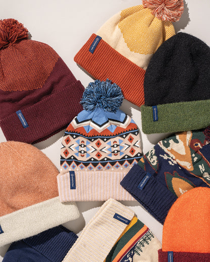 Recycled Chevron Pom Beanie (Flat-lay): Multiple knit beanies arranged together, center one features geometric pattern with blue pompom