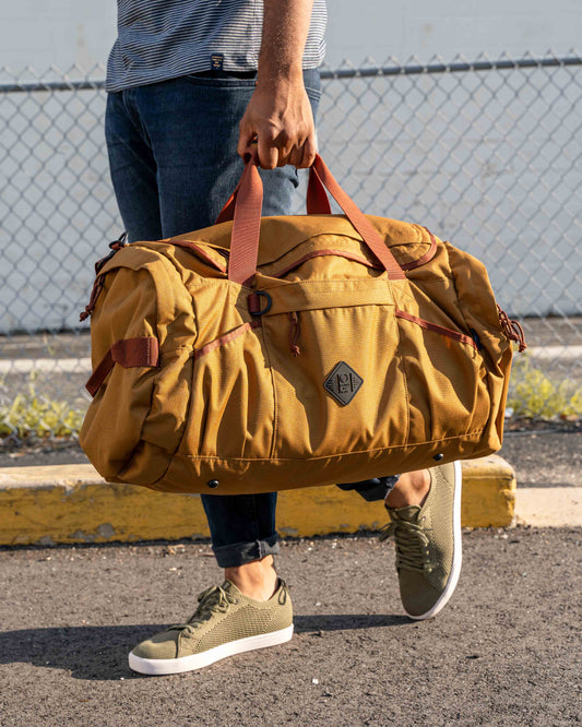 Camel (R)evolution™ 55L Carry-On Duffle (In-use): Person carrying large tan duffle with rust-colored handles in parking lot