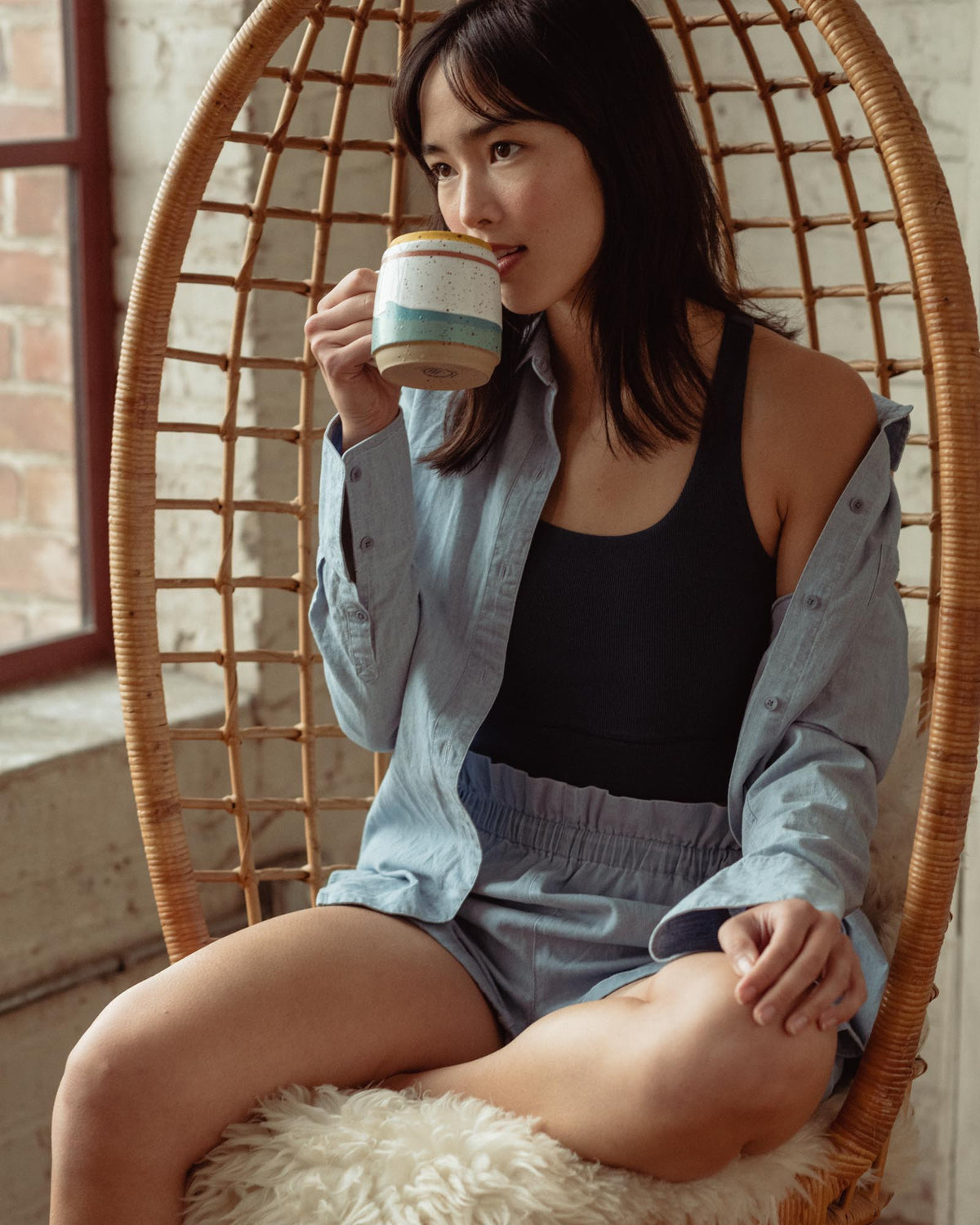 Eggshell 16 oz. Stoneware Mug (In-use): Woman sipping from speckled mug with pastel stripe bands, seated in rattan chair