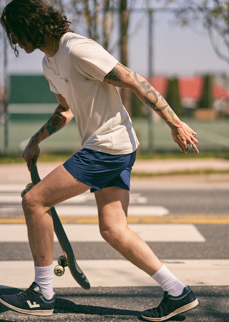 Estate Blue Recycled Sport Short (In-use): Tattooed skater wearing navy blue shorts crosses a sunny street