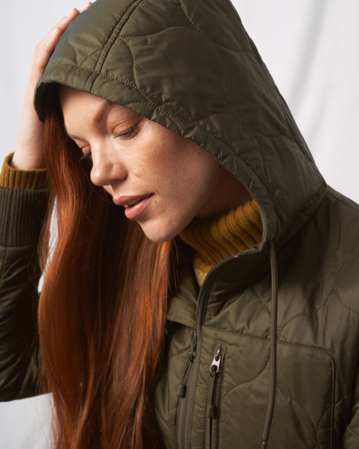 Forest Floor Bison Ultralight Long Liner Jacket (In-use): Woman with red hair wearing olive quilted hooded jacket, eyes closed