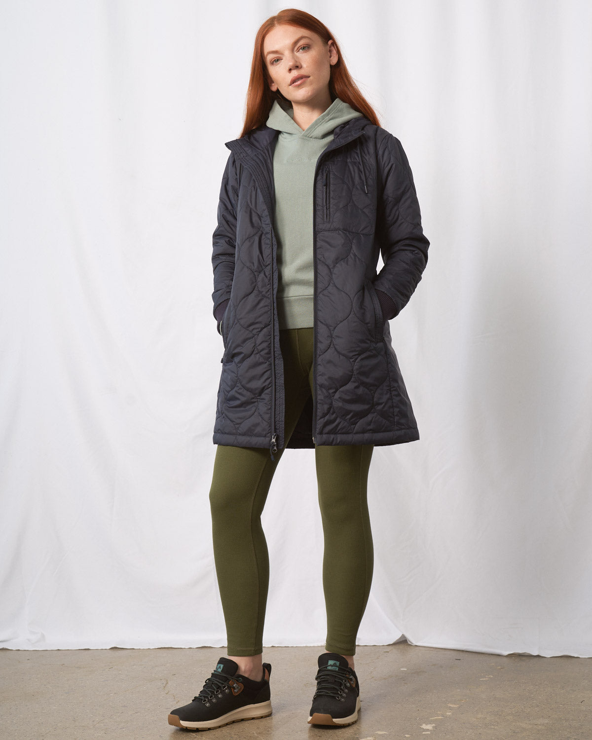 Night Sky Bison Ultralight Long Liner Jacket (In-use): Woman wearing open dark navy quilted long coat, olive leggings, sage hoodie