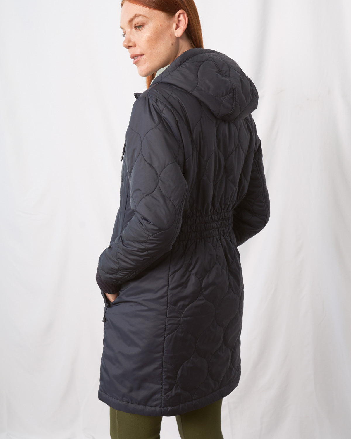 Night Sky Bison Ultralight Long Liner Jacket (Back): Dark navy quilted hooded long jacket with elastic waist, worn by woman