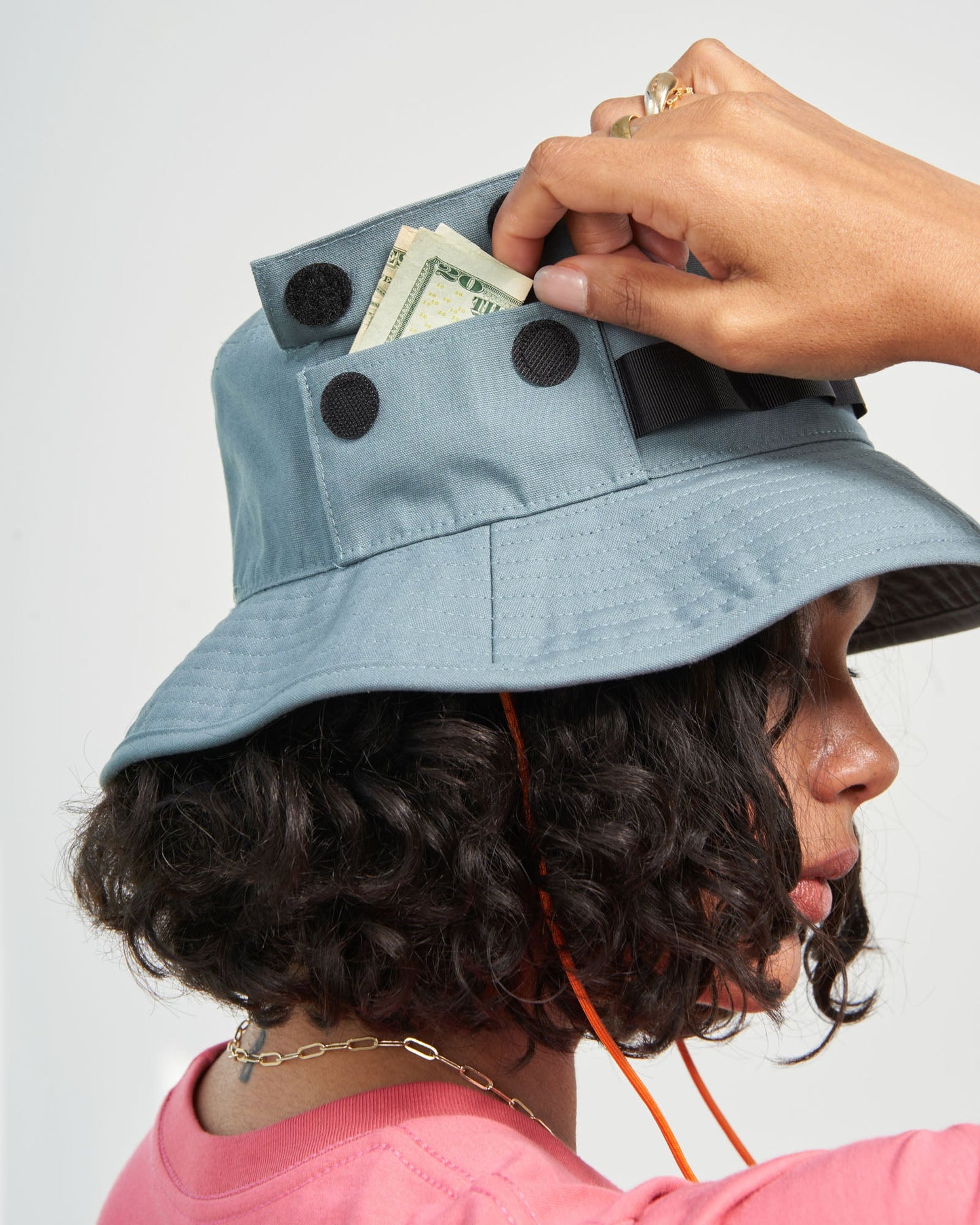 Sky Blue Organic Bucket Stash Hat (In-use): Person retrieving folded bills from a velcro-closure pocket on a slate-blue bucket hat