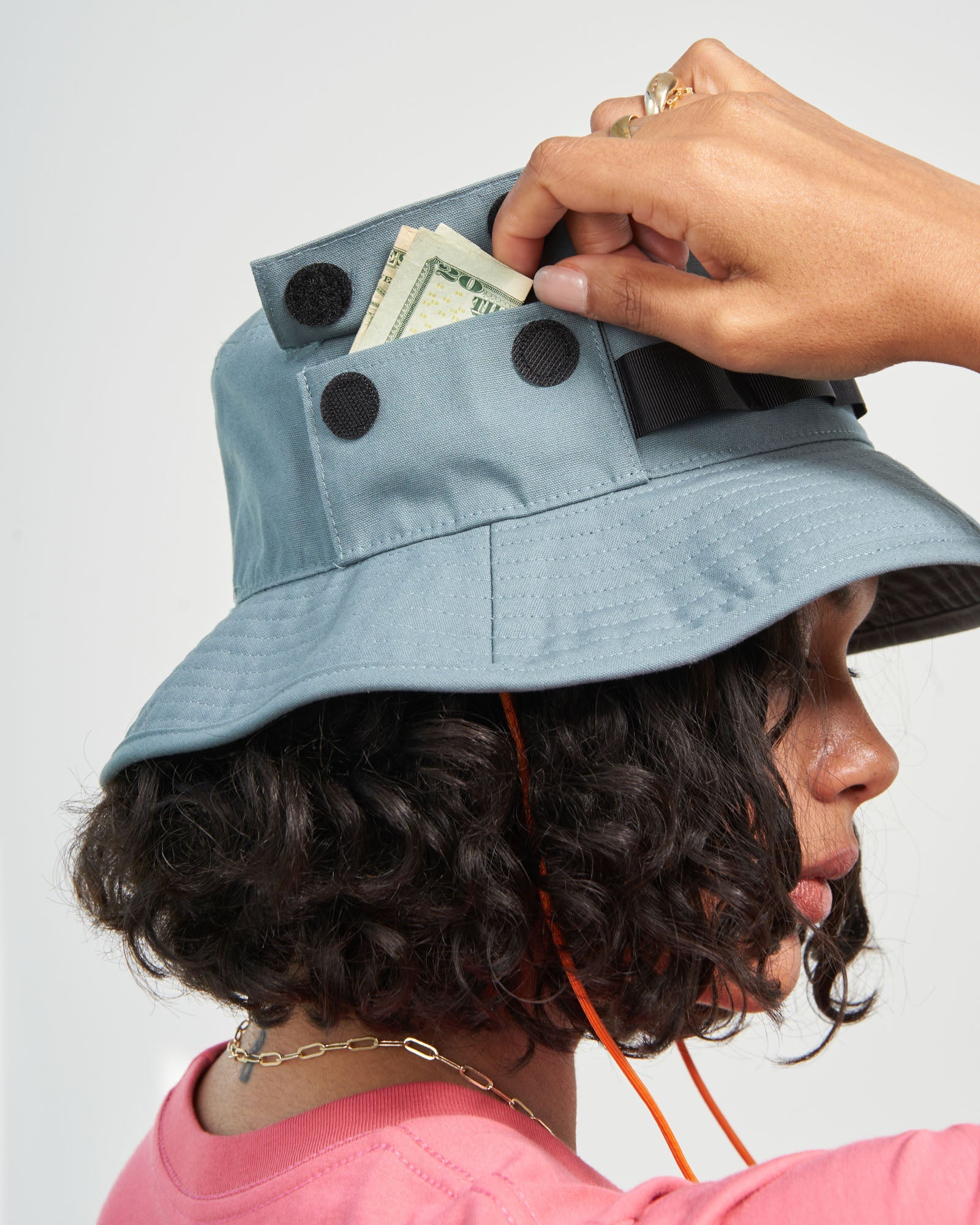 Sky Blue Organic Bucket Stash Hat (In-use): Person retrieving folded bills from a velcro-closure pocket on a slate-blue bucket hat