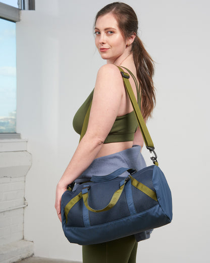 Blueberry (Re)active™ 16L Mini Duffle (In-use): Woman in olive sports bra carries navy blue duffle with olive straps