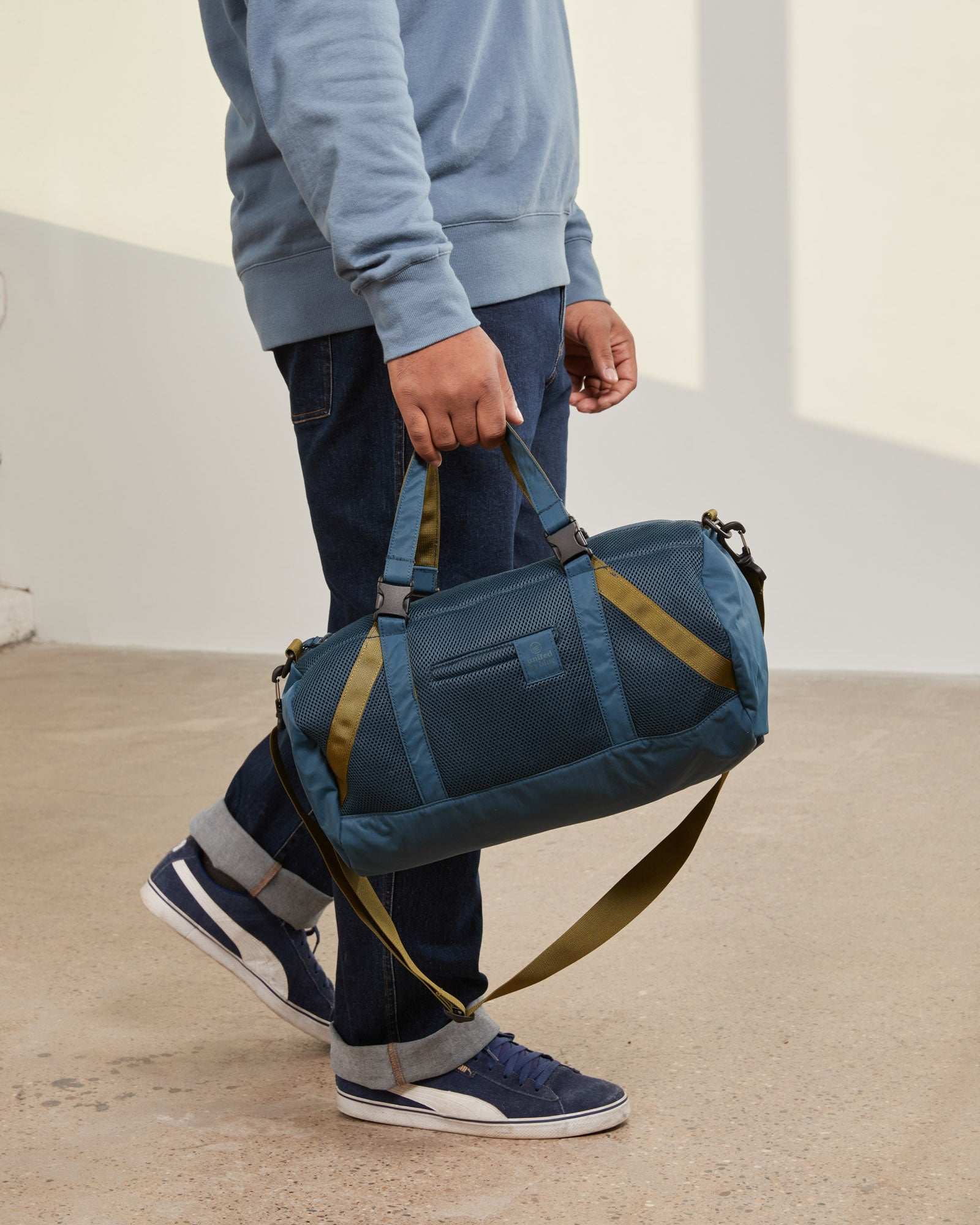 Blueberry (Re)active™ 16L Mini Duffle (In-use): Person carrying teal duffle with olive straps, mesh panels, and shoulder strap