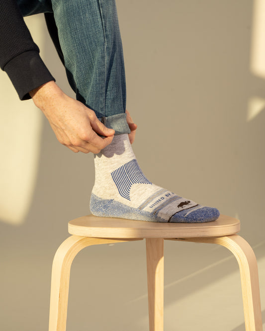 Blue Bison Wool Trail Sock (In-use): Person pulling up gray and blue ankle sock, foot resting on wooden stool